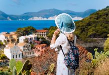 Must Have Clothing Items When Travelling To Greece Tourist woman with blue sunhat, white clothes and travel backpack admire view of colorful tranquil village Assos on sunny day. Visiting Kefalonia during summer time on Greece travel vacation