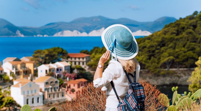Must Have Clothing Items When Travelling To Greece Tourist woman with blue sunhat, white clothes and travel backpack admire view of colorful tranquil village Assos on sunny day. Visiting Kefalonia during summer time on Greece travel vacation
